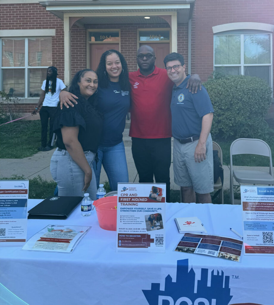 PCSI staff standing with the mayor of Pittsburgh behind an outreach table at a National Night Out event at Bedford Dwellings, with flyers and program information displayed on the table.