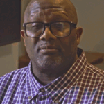 Shawn is a black man, bald and wearing eyeglasses and a purple plaid button down shirt.
