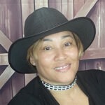Photo of Nikia, a Black woman wearing a black blouse and a black hat with a brim.